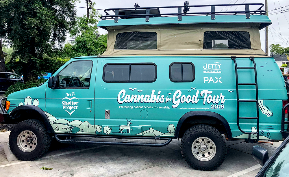 A teal Jetty Shelter Project van from the 2019 “Cannabis for Good Tour,” featuring mountain and forest graphics, the Jetty and PAX logos, and the slogan “Protecting patient access to cannabis,” representing Jetty’s advocacy for compassionate care through the Shelter Project.