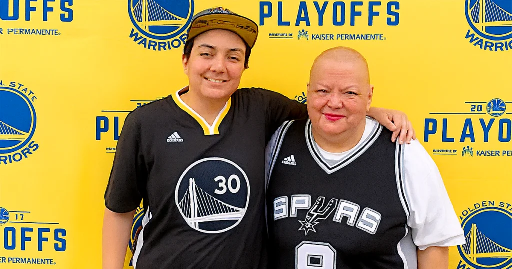 Rachel Gaeta and Amanda Gaeta smile together in front of a Golden State Warriors playoff banner, representing strength and positivity in her mothers journey with medical marijuana for cancer through the Jetty Shelter Project.