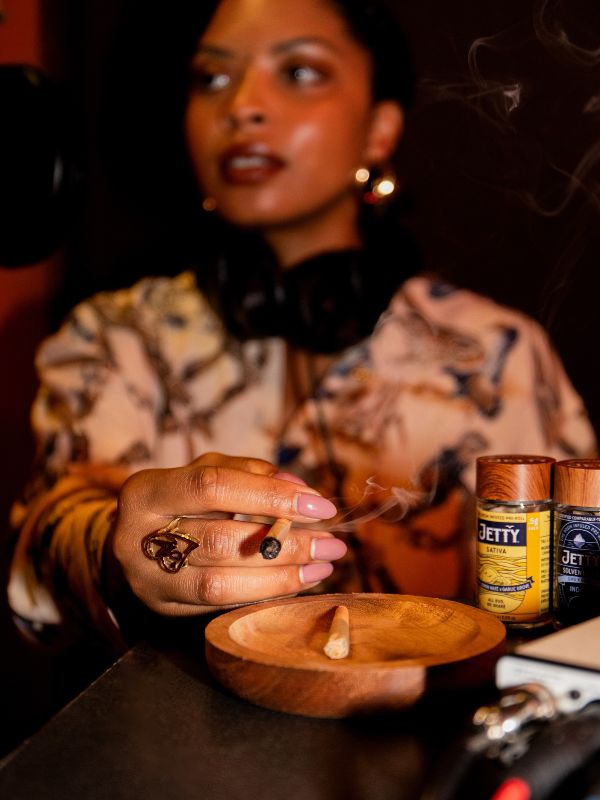 close up up a woman's hand with a pre-roll joint burning into a wood ashtray and jetty infused pre-roll jars are next to it.