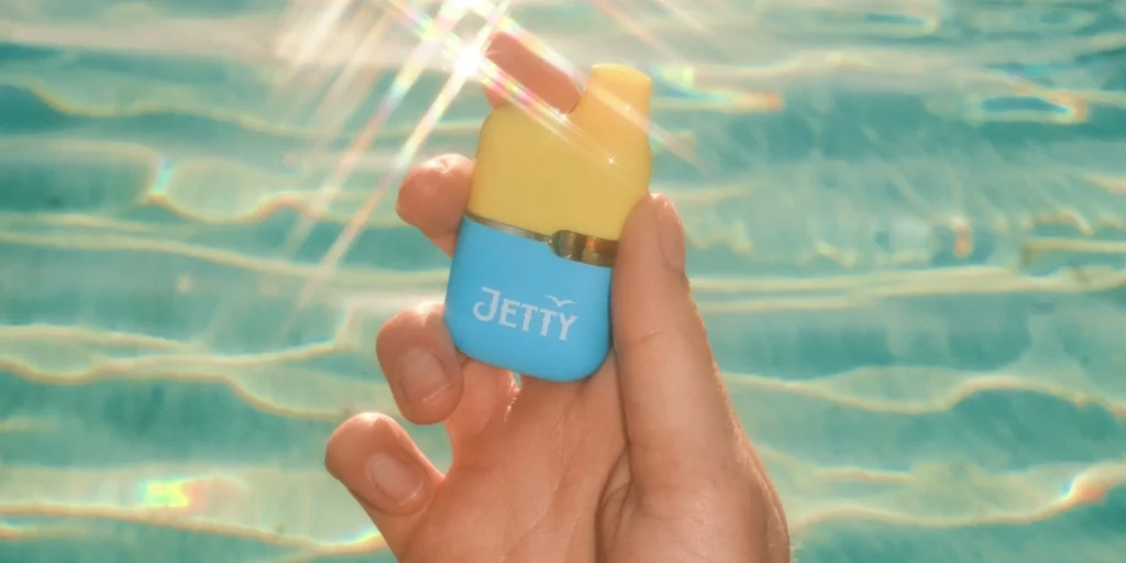 Hand holding a small Jetty CCELL Tank with a yellow mouthpiece and blue base, positioned in front of sparkling turquoise pool water in bright sunlight.