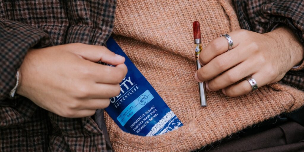 Person wearing a brown plaid jacket and tan sweater holds a silver solventless 510 thread vape cart with a red mouthpiece, while a blue Jetty Solventless cannabis product package labeled “Grape Gas” is tucked into their sweater pocket.
