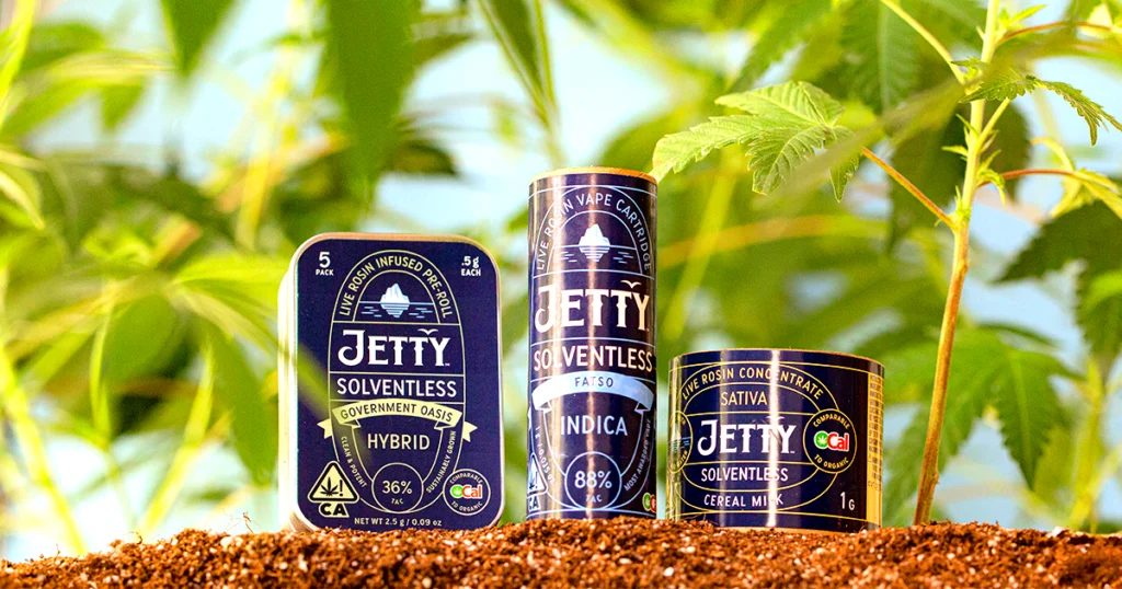 Three Jetty Solventless cannabis products—pre-rolls, vape cartridge, and concentrate—are displayed in soil with green cannabis leaves in the background, highlighting the brand’s natural, live rosin-based extraction process.