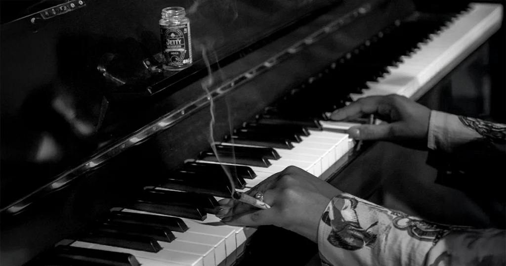 In a black-and-white shot, a person plays piano while holding a lit Jetty infused pre-roll, with a Jetty Solventless Indica jar resting on the piano ledge, capturing the harmony between music, creativity, and Jetty’s premium cannabis craftsmanship.