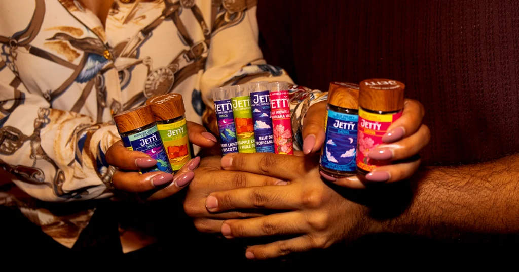 Two people holding multiple Jetty High-THC infused pre-roll jars and tubes together in warm retro lighting, showcasing colorful strain labels from Jetty’s Greatest Hits collection.