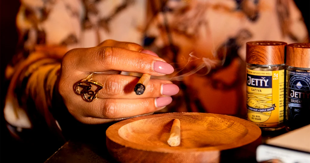 A woman with polished nails and gold jewelry holds a lit Jetty Sativa High-THC infused pre-roll beside an ashtray, with Jetty Sativa and Indica jars in the background, emphasizing the flavor, quality, and craftsmanship of Jetty’s infused joints.