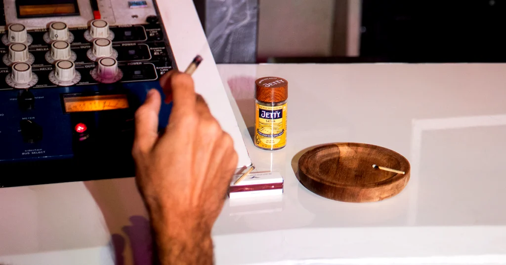 A person sits in a music studio holding a lit pre-roll near an audio mixing console, with a Jetty Sativa High-THC infused pre-roll jar, a matchbook, and a wooden ashtray on the table, illustrating the creative process behind Jetty’s handcrafted cannabis products.