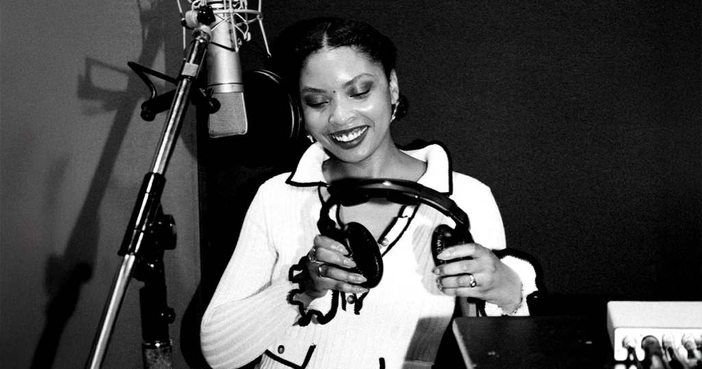 A woman smiles in a recording studio, holding headphones in front of a professional microphone setup, symbolizing the artistry, focus, and creativity that inspire Jetty’s premium infused pre-rolls and the Greatest Hits series.