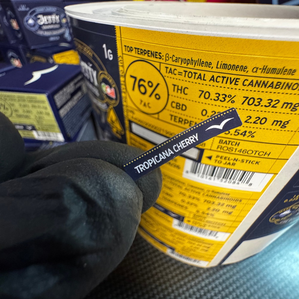 Close up of a black gloved hand holding a tiny peel-n-stick vape cart label reading tropicana Cherry.