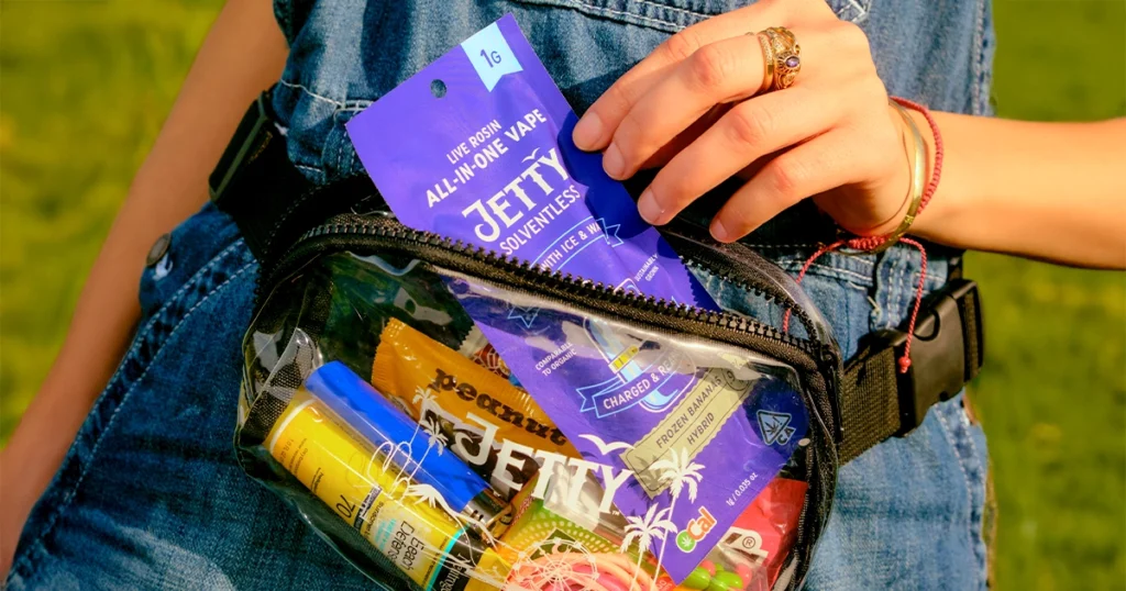 Person wearing denim overalls outdoors holding a Jetty Solventless Live Rosin All-In-One Vape 1g package in a clear waist bag filled with Jetty products, including Frozen Banana Hybrid vape, sunscreen, and colorful candy accessories, under warm sunlight.