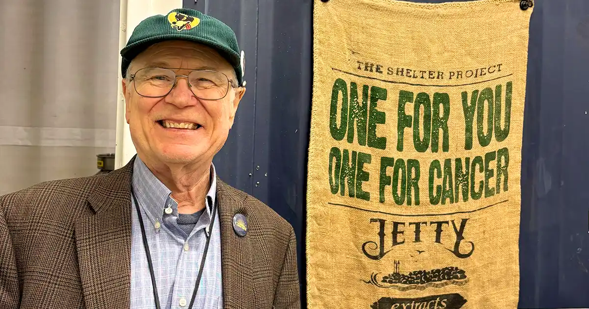 Jim Stockinger smiling in California, standing beside a Shelter Project banner reading “One for You, One for Cancer” by Jetty Extracts.