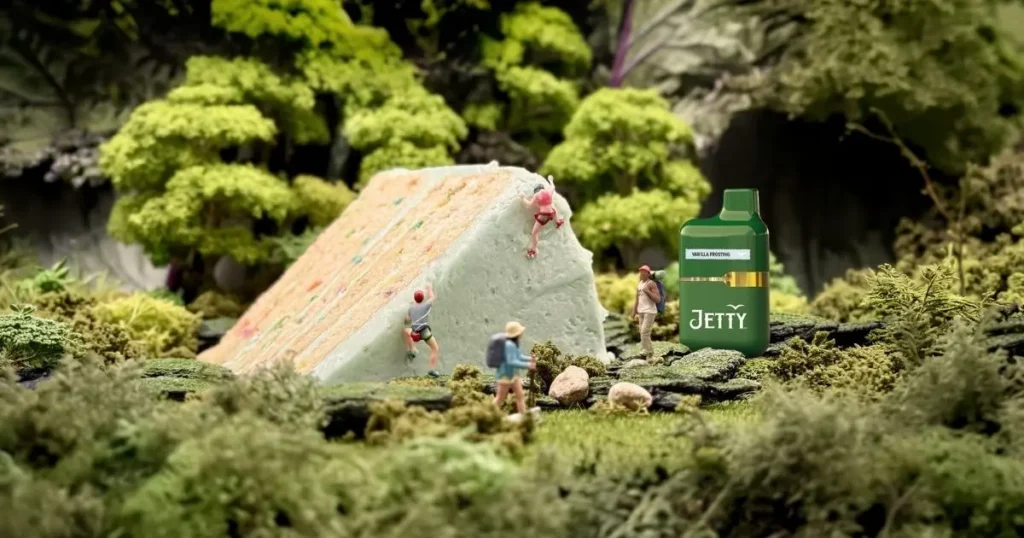 Miniature hikers climb a frosted cake slice beside a green Jetty Vanilla Frosting vape in a mossy forest.
