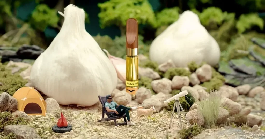 Miniature camper relaxes beside a tent and telescope near a Jetty Garlic Mac vape surrounded by oversized garlic bulbs.